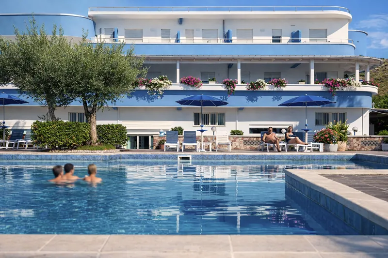 Piscina con ospiti in acqua davanti alla facciata bianca e blu dell'hotel